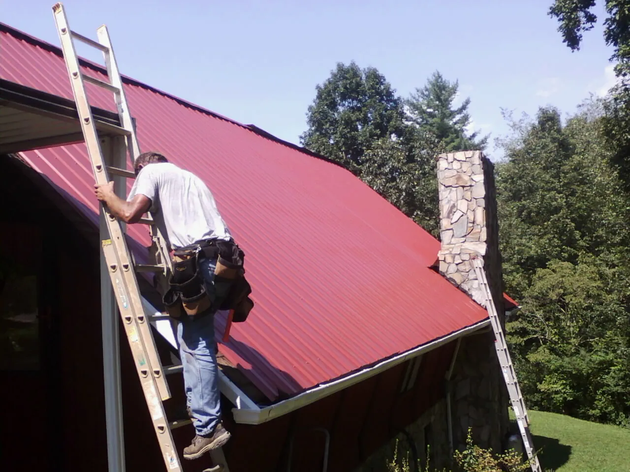 Roofer on a ladder beside a red metal roof and stone chimney in Billerica